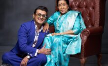 Sonu Nigam in blue suit sitting on the ground next to Asha Bhosle in cyan blue sari on a chair