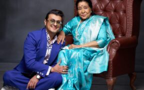 Sonu Nigam in blue suit sitting on the ground next to Asha Bhosle in cyan blue sari on a chair