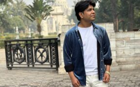 Ankit Tiwari wearing blue jacket over white t-shirt and trousers, looking up with a white dome structure in the background