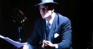 Ash Chandler seated on a white box wearing black suit and hat