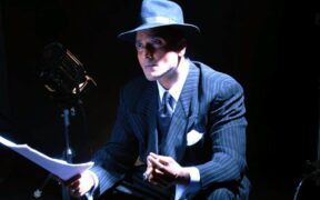 Ash Chandler seated on a white box wearing black suit and hat