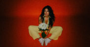 Tiana Tara wearing yellow dress, holding white flowers and sitting in red background