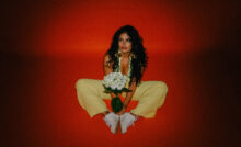 Tiana Tara wearing yellow dress, holding white flowers and sitting in red background