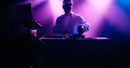 A young African American deejay performs for a crowd at a city night club. Colorful stage lights illuminate the stage behind him.